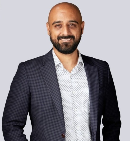 Smiling bald man with beard wearing a navy checked blazer and patterned shirt against a grey background