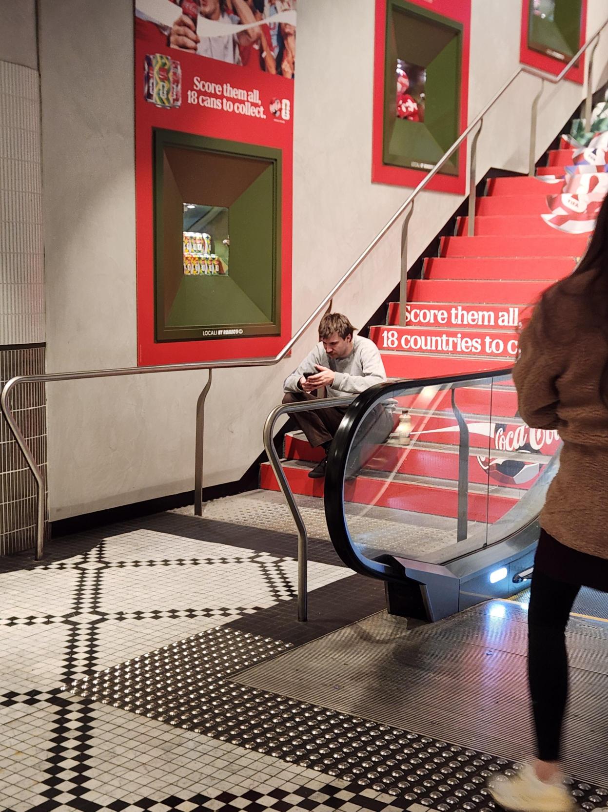 Man sitting on red Coca-Cola branded stadium stairs looking at phone, next to escalator in public venue.