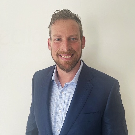 Professional headshot of a smiling man with beard wearing a navy blazer and light blue checked shirt against white background