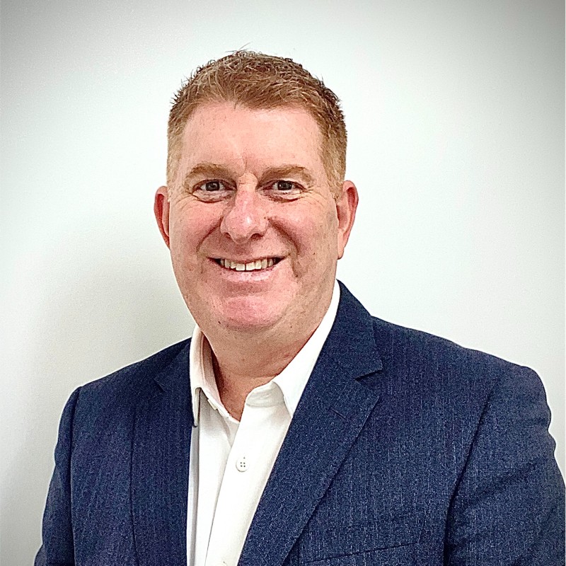 Smiling middle-aged man with short red hair wearing a navy blazer and white shirt against a light background.