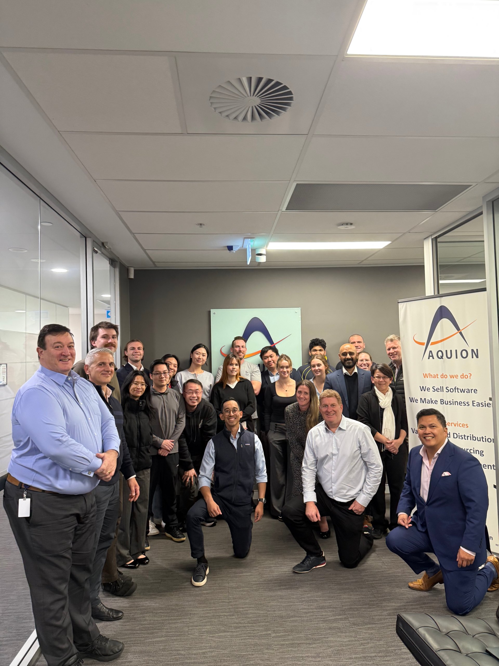 Aquion team group photo in office, gathered in front of company logo and branded banner, smiling together.